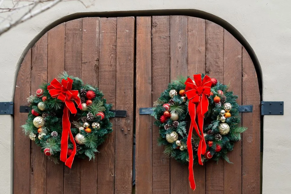 Wooden doors with large Christmas wreaths, adorned with red ribbons and festive ornaments, are displayed.