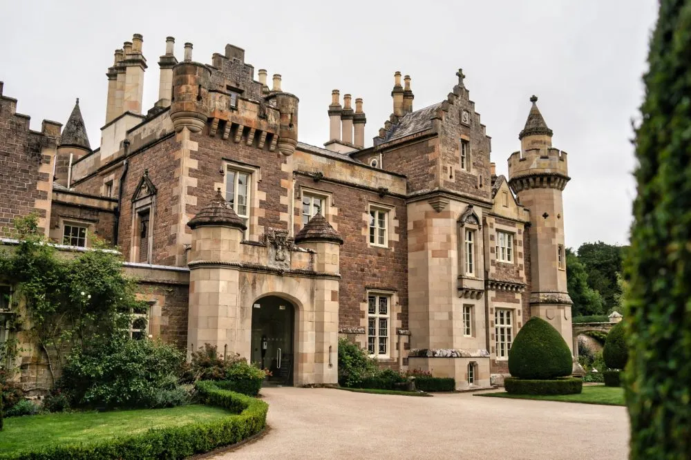 Historic castle with turrets and stone facade, surrounded by manicured lawns and hedges under a cloudy sky.