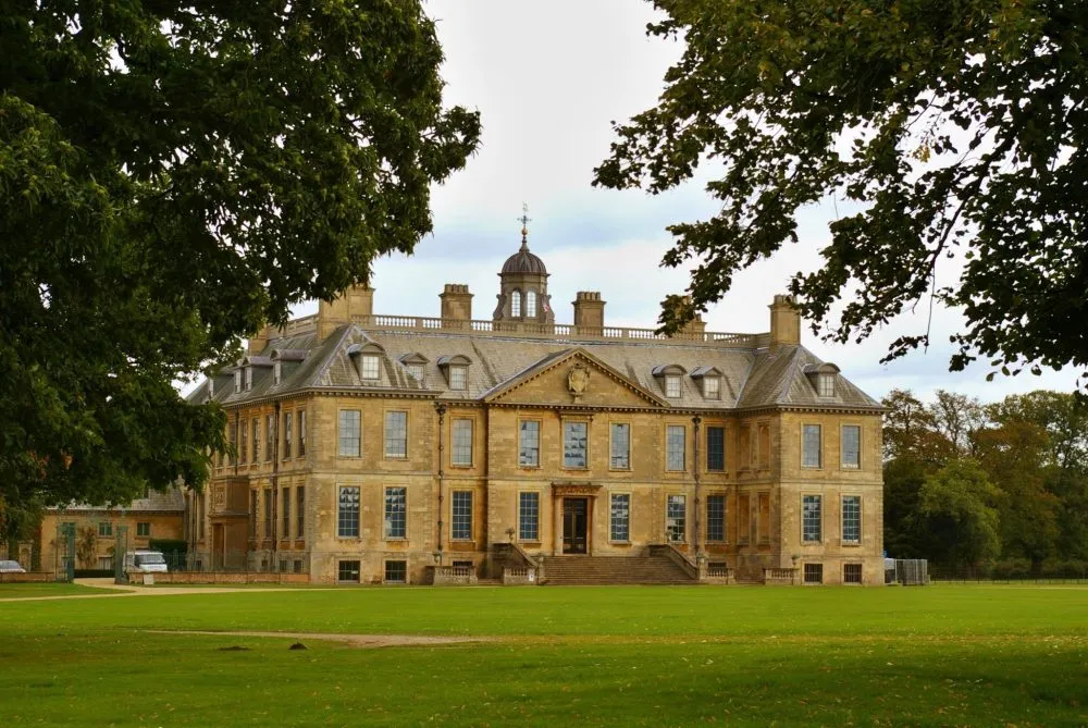 A large, historic mansion with multiple chimneys and a central cupola, surrounded by trees and a vast grassy area.