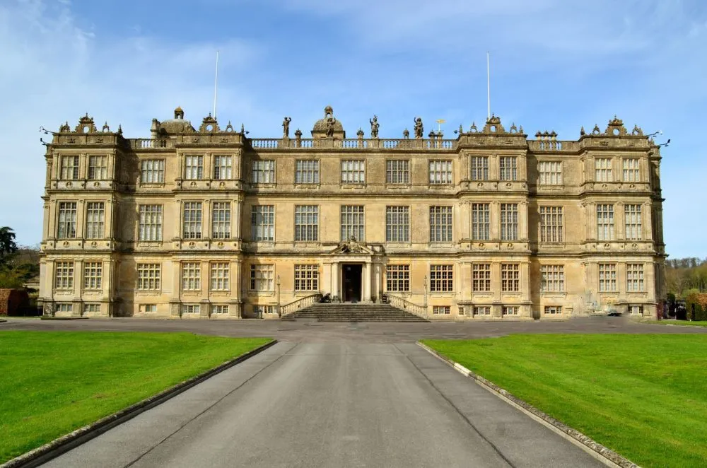 Grand historic mansion with symmetrical facade, ornate sculptures, and expansive lawn under a clear blue sky.